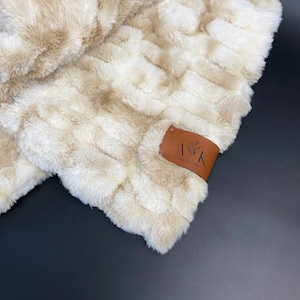 May include: A white and beige faux fur throw blanket with a brown leather tag that says "A&K July 27, 2020".
