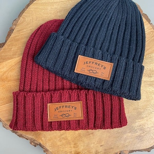 May include: Two ribbed knit beanies, one in navy blue and the other in burgundy, are displayed on a wooden surface. Each beanie has a brown leather patch with the text "JEFFREY'S GROCERY" and a logo.
