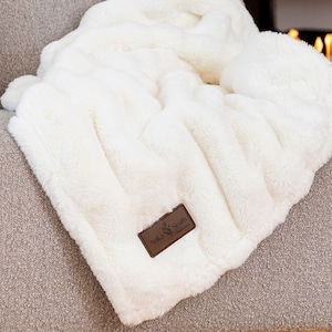 May include: A white faux fur throw blanket with a brown leather tag that says "Arka Smith" on a beige armchair in front of a fireplace.