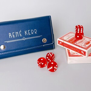 May include: A navy blue leatherette case with the name "RENÉ KERR" in white lettering, silver snaps, and a white stitched border. Beside it are three red dice and two decks of playing cards.
