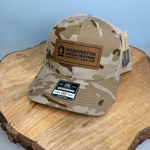 May include: A camouflage-patterned trucker hat featuring a rectangular brown leather patch with the black text "WASHINGTON WATER HEATERS HEATING AND AIR" and a logo. A black and white sticker on the brim reads "RICHARDSON 112 TRUCKER".