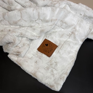 May include: A soft white and light beige faux fur throw blanket featuring a textured, wavy pattern. A brown, diamond-shaped leather patch is attached to the corner, engraved with the letters A and D, a heart symbol, and the date June 24, 2019.