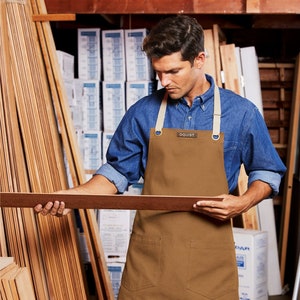 May include: A person wearing a brown canvas apron with the word "OQUIST" on the front pocket, holding a piece of wood in a lumber yard.