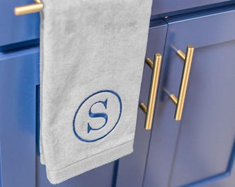 May include: A light gray hand towel featuring a dark blue embroidered letter S inside a circular border. The towel hangs from a brushed gold metal handle attached to a vibrant blue cabinet door with matching gold hardware.