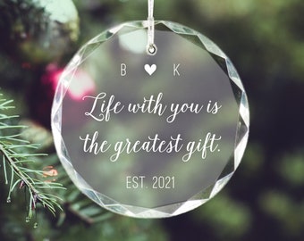 May include: Clear, faceted glass Christmas ornament with the text "Life with you is the greatest gift." and "EST. 2021." The ornament is hanging on a Christmas tree. The initials "B" and "K" are above the text, separated by a heart symbol.