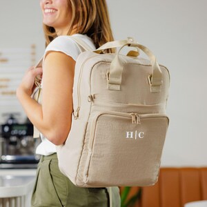 May include: A beige backpack with a top handle, shoulder straps, and a front zippered pocket. The backpack has the initials "H|C" in white on the front. The person in the image is wearing a white shirt and green pants.
