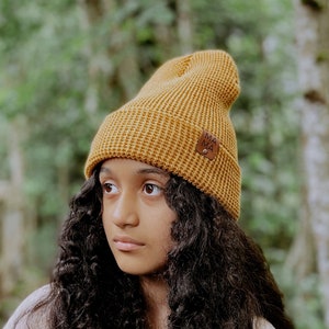 May include: A mustard yellow waffle-knit beanie with a folded cuff. A small, square, brown faux-leather patch is attached to the side of the cuff, featuring the word MAMA embossed in black capital letters and secured with a single silver metal rivet.