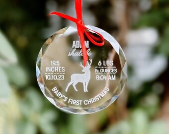 May include: A clear, faceted glass Christmas ornament with a red ribbon. The ornament features engraved text: baby's first Christmas, a deer silhouette, and baby's birth details: 19.5 inches, 6 lbs 14 ounces, and the date and time of birth.