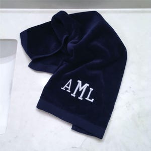 Puede incluir: Una toalla de mano de color azul marino profundo con una textura suave y afelpada y un dobladillo acabado. El centro de la toalla está bordado con las letras mayúsculas blancas AML en una fuente serif clásica, creando un contraste claro.