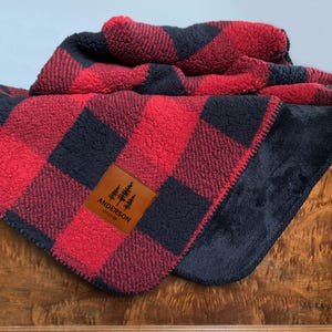 May include: A plush, textured blanket featuring a red and black buffalo plaid pattern. A rectangular brown faux leather patch is stitched onto the fabric, displaying the text "ANDERSON" above three pine tree icons and the date "07.27.89".