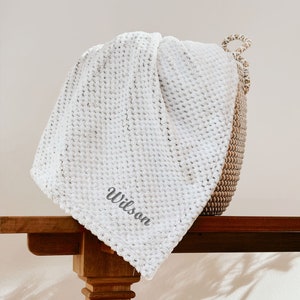 May include: White waffle knit throw blanket with the name "Wilson" embroidered in gray on the corner. The blanket is draped over a wooden table.