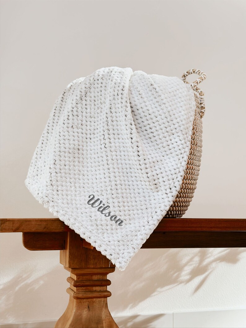 May include: White waffle knit throw blanket with the name "Wilson" embroidered in gray on the corner. The blanket is draped over a wooden table.