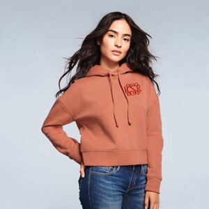 May include: A cropped rust-colored hoodie with a drawstring hood and a red embroidered monogram on the chest. The hoodie is paired with blue jeans. The model is wearing the hoodie.