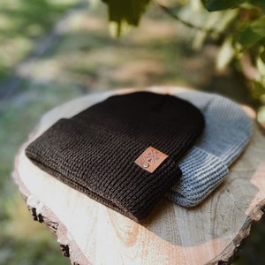May include: Two knit beanies, one black and one gray, stacked on a wooden slice. The black beanie has a brown leather patch with a logo on it.