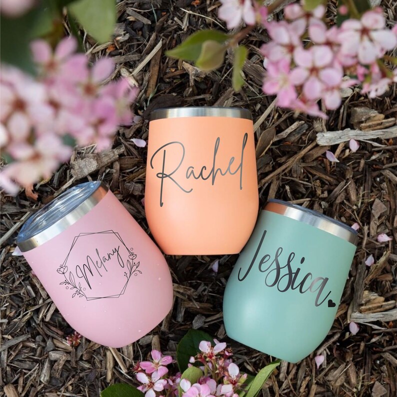 May include: Three stainless steel stemless tumblers with lids in pastel pink, peach, and mint green. The pink tumbler features the name Melany inside a floral hexagon. The peach tumbler displays the name Rachel, and the mint tumbler shows the name Jessica.