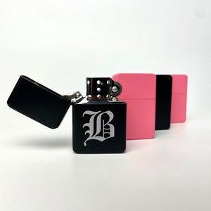 May include: A black lighter with a silver hinge and a white letter "B" engraved on the front. The lighter is next to two pink and black lighters.