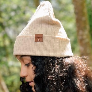 May include: A beige ribbed knit beanie featuring a folded cuff and a small square brown leather patch on the front. The patch is embossed with the text Karmin and is secured with a single metallic rivet at the bottom center of the square label.