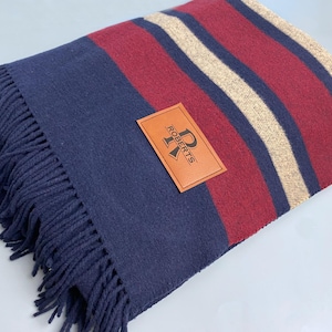 May include: A folded navy blue blanket with burgundy and tan stripes. The blanket has a navy fringe along one edge and a brown leather tag with the name "ROBERTS" on it.