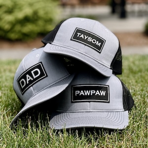 May include: Three gray and black trucker hats with black patches. The patches have white text that reads "Taysom", "Dad", and "Pawpaw".