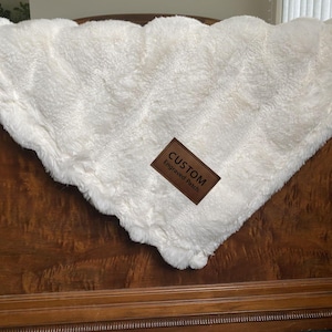 May include: A fluffy, off-white blanket with a scalloped edge. A brown leather patch with the words "CUSTOM Engraved Patch" is sewn onto the blanket. The blanket is folded over a wooden surface.