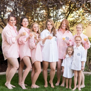 May include: A group of eight people wearing pink and white button-down shirts, some holding glasses of champagne. The people are smiling and looking at the camera. The image is taken outdoors in front of a green bush.