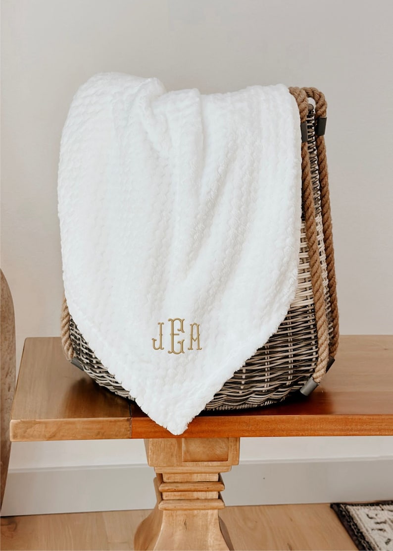 May include: A white plush throw blanket with a monogrammed "J.E.M." in gold thread, folded and placed in a wicker basket with rope handles.