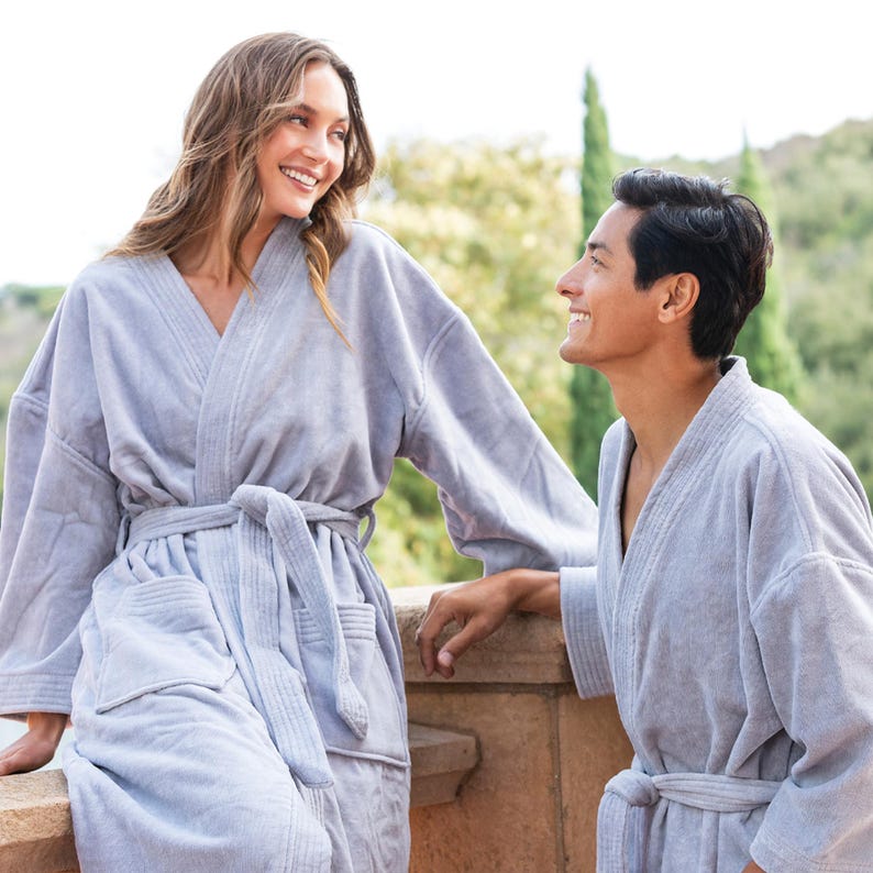 May include: Two people wearing matching gray bathrobes stand on a balcony and look at each other. The robes have a waffle texture and are tied at the waist.