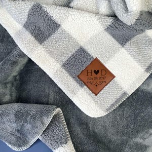 May include: A plush throw blanket featuring a grey and white buffalo check pattern on one side and solid grey on the other. A diamond-shaped brown leather patch is stitched to the corner, displaying the text H heart D and July 28, 2017.