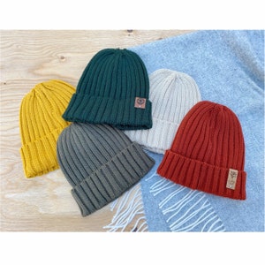 May include: Five knitted beanies in different colors: mustard yellow, forest green, olive green, beige, and rust orange. Each beanie has a small leather patch with a logo on the side.