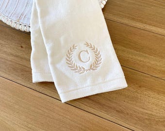 May include: A white hand towel featuring an embroidered beige laurel wreath design centered around the letter C. The towel has a textured fabric finish and a hemmed edge at the bottom, showcasing a clean and simple aesthetic for bathroom decor.