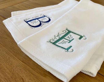 May include: Two white cotton towels featuring embroidered monograms. One towel displays a dark blue letter B with decorative flourishes, while the other towel features a teal green letter E with similar intricate leaf-like embroidery details.