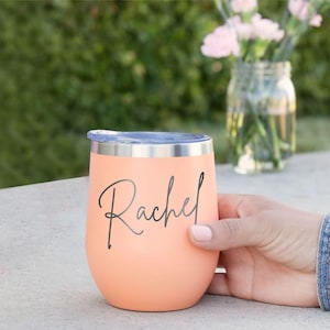 May include: A peach-colored stainless steel tumbler with a metallic silver rim and lid. The name Rachel is printed in a dark gray cursive script on the front of the cup. The tumbler has a rounded, egg-shaped body and a smooth matte finish.