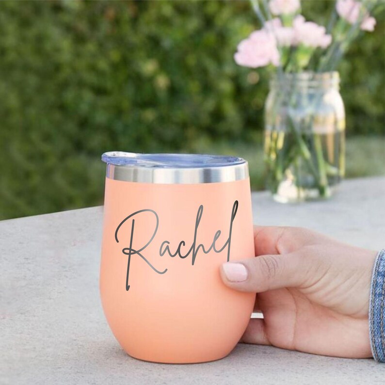 May include: A peach-colored stainless steel tumbler with a metallic silver rim and lid. The name Rachel is printed in a dark gray cursive script on the front of the cup. The tumbler has a rounded, egg-shaped body and a smooth matte finish.