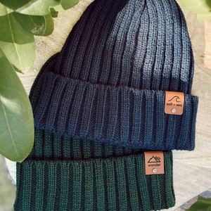 May include: Two knitted beanies, one dark blue and one dark green, with leather patches. The blue beanie has a wave logo and the text "salt + sea". The green beanie has a mountain logo and the text "wander".