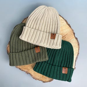May include: Three ribbed knit beanies in cream, olive green, and dark forest green. Each hat features a folded cuff and a small, rectangular brown leather-like patch attached with a metal rivet, embossed with a heart and the text CHOSEN FAMILY.