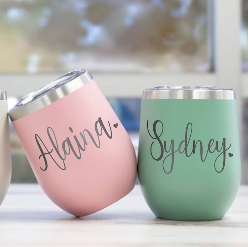 May include: Two stainless steel wine tumblers with personalized names. The tumbler on the left is pink with the name "Alaina" and a small heart. The tumbler on the right is green with the name "Sydney" and a small heart.