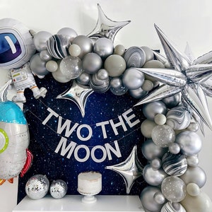 K&ouml;nnte beinhalten: Dekoration f&uuml;r eine Party zum Thema Weltraum mit einem Astronauten- und Raketenballon, einem Ballonbogen und einem Banner mit der Aufschrift "TWO THE MOON". Das Farbschema umfasst silberne, graue und wei&szlig;e Ballons mit einem dunkelblauen Hintergrund.