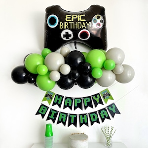 Könnte beinhalten: Ein schwarzes und grünes Geburtstagsdekorationsset zum Thema Videospiele. Das Set enthält einen großen schwarzen und grünen aufblasbaren Videospiel-Controller mit dem Text "Epic Birthday!" darauf, ein schwarzes und grünes Banner mit der Aufschrift "Happy Birthday" und eine Girlande aus schwarzen, grünen und grauen Ballons.