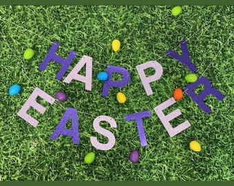May include: Purple glitter letters spelling "Happy Easter" on a green grass background with colorful Easter eggs scattered around.