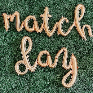 May include: Gold foil letter balloons spelling out "match day" on a green artificial grass background.