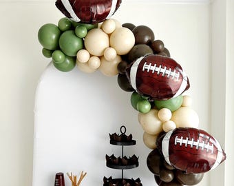 Puede incluir: Un arco de globos marrones y verdes con globos con forma de balón de fútbol y un soporte para cupcakes negro de tres niveles con cupcakes marrones.
