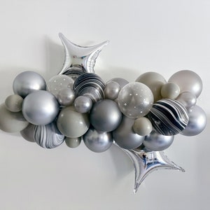 May include: A cluster of gray and silver balloons, including star-shaped foil balloons, arranged in a decorative display. Some balloons have a marbled effect, while others feature small white stars. The balloons vary in size and shade.