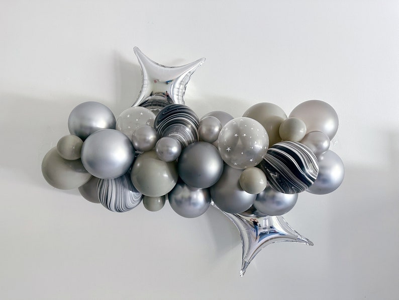 May include: A cluster of gray and silver balloons, including star-shaped foil balloons, arranged in a decorative display. Some balloons have a marbled effect, while others feature small white stars. The balloons vary in size and shade.