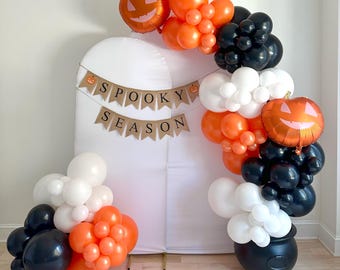 May include: A Halloween-themed balloon arch featuring orange, black, and white balloons. Two pumpkin-shaped foil balloons and a banner that reads "Spooky Season" are included. The arch is set against a white backdrop, with a black cauldron at the base.