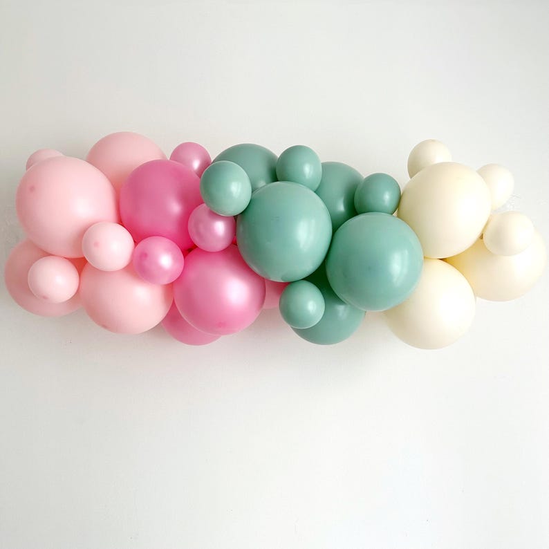 May include: A garland of pastel pink, sage green, and ivory balloons. The balloons are arranged in a cascading pattern, creating a festive and whimsical look.