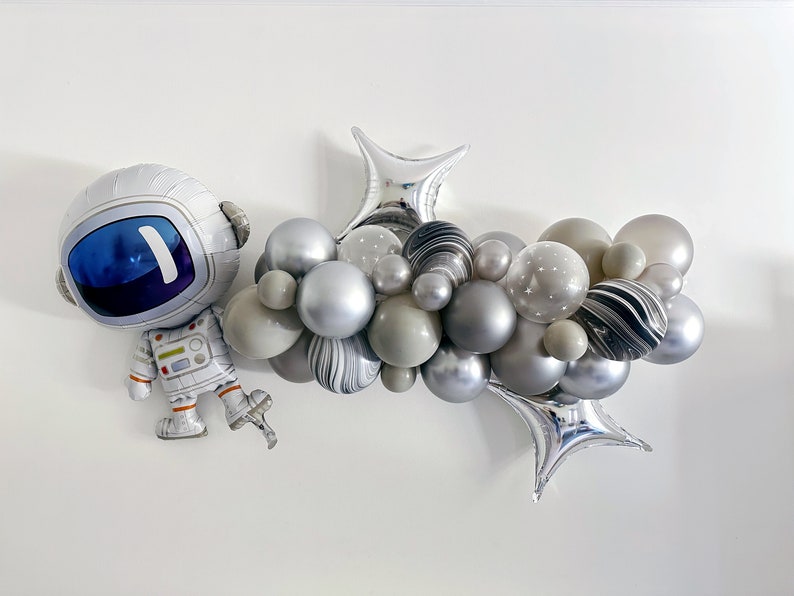May include: A silver astronaut-shaped balloon and a cluster of gray, silver, and marble-patterned balloons. The balloon arrangement includes star-printed balloons and star-shaped foil balloons, creating a space-themed decoration.