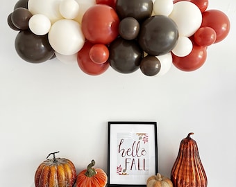 May include: A seasonal display with a balloon garland in white, brown, and rust shades above a shelf. The shelf features orange and brown decorative pumpkins, a framed print saying "hello FALL", and a small woven pumpkin.