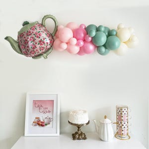 May include: A tea party themed balloon arch with pastel pink, green, and cream balloons. A teapot balloon is attached to the left side of the arch. A framed print with the text "tea for two" is on a table with a cake stand, a white teapot, and a stack of teacups.