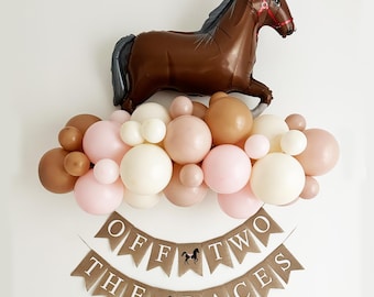 May include: A brown horse balloon with a red bridle, surrounded by a garland of pink, beige, and white balloons. A burlap banner with the text "Off to the Races" is below the balloons.