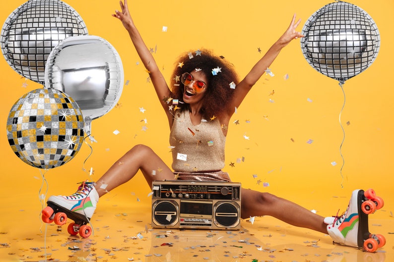 May include: A woman wearing roller skates and sunglasses is sitting in front of a boombox with silver disco ball balloons and confetti around her.